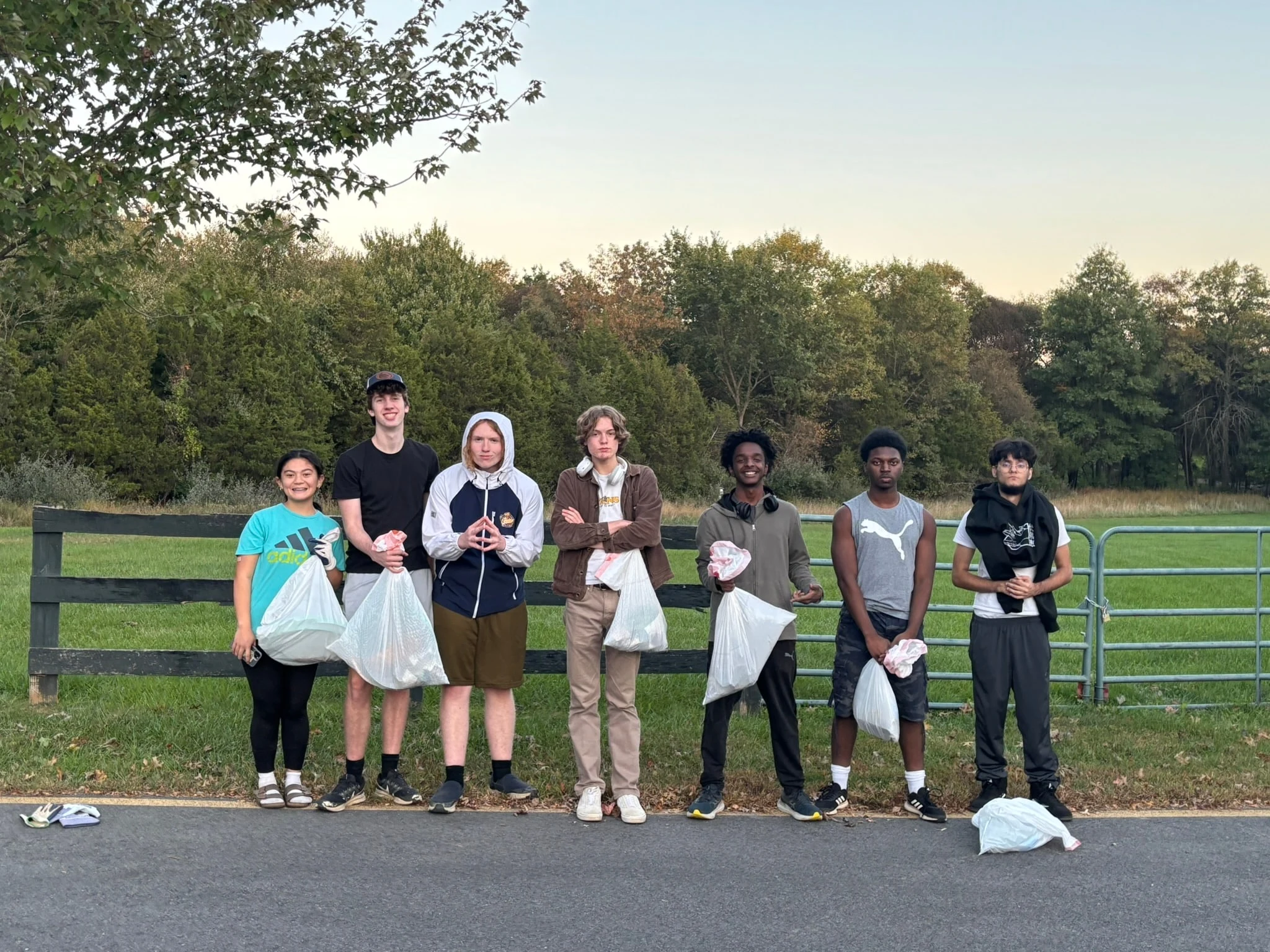 Volunteers holding bags of trash in a row along a black fence beside a country road at dusk
