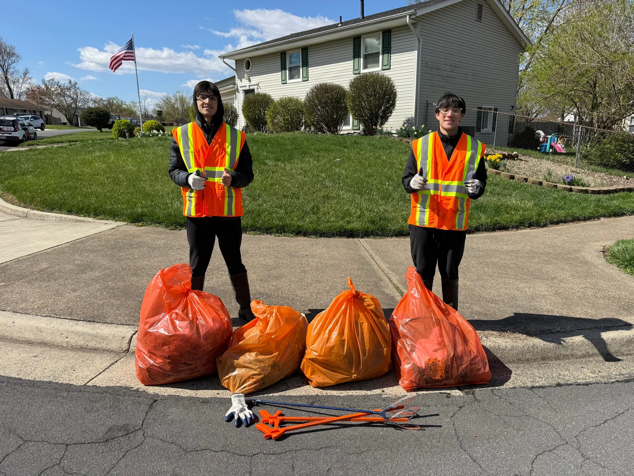 Two volunteers in orange safety vests standing behind four orange trash bags at the edge of a residential street